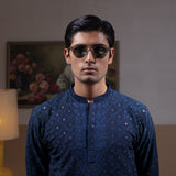 Man wearing Arsalan Iqbal's Navy Blue Allover Sequin Embroidered Kurta with dark navy trousers, black loafers and sunglasses, posed against a beige backdrop with yellow lamp and floral painting