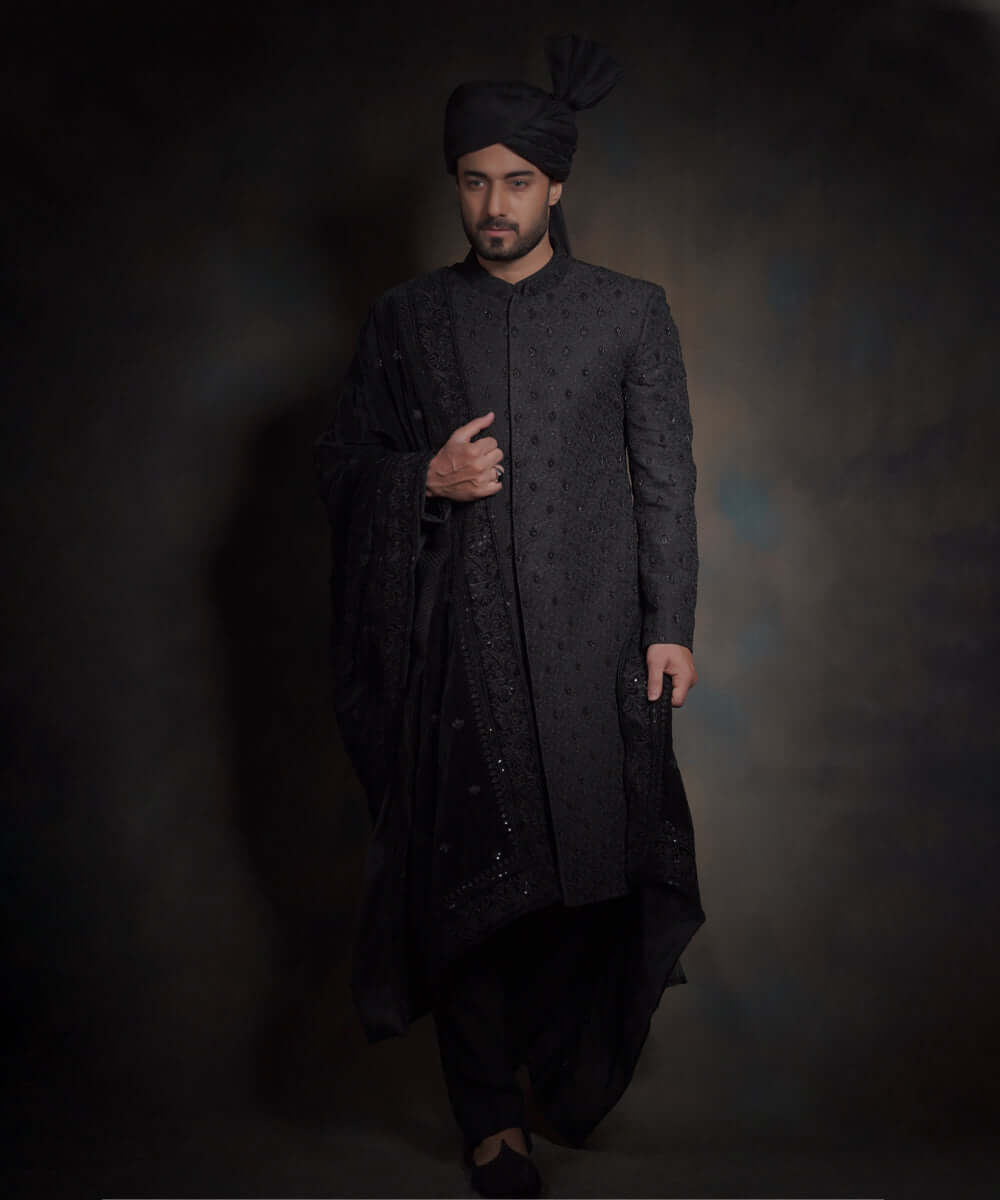 Arsalan Iqbal midnight black luxury sherwani for men, featuring spaced-out tonal floral motifs, a matching black turban with a high pleat, a draped velvet shawl, and black trousers.