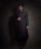 Arsalan Iqbal midnight black luxury sherwani for men, featuring spaced-out tonal floral motifs, a matching black turban with a high pleat, a draped velvet shawl, and black trousers.