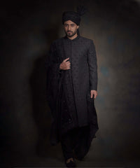 Arsalan Iqbal midnight black luxury sherwani for men, featuring spaced-out tonal floral motifs, a matching black turban with a high pleat, a draped velvet shawl, and black trousers.