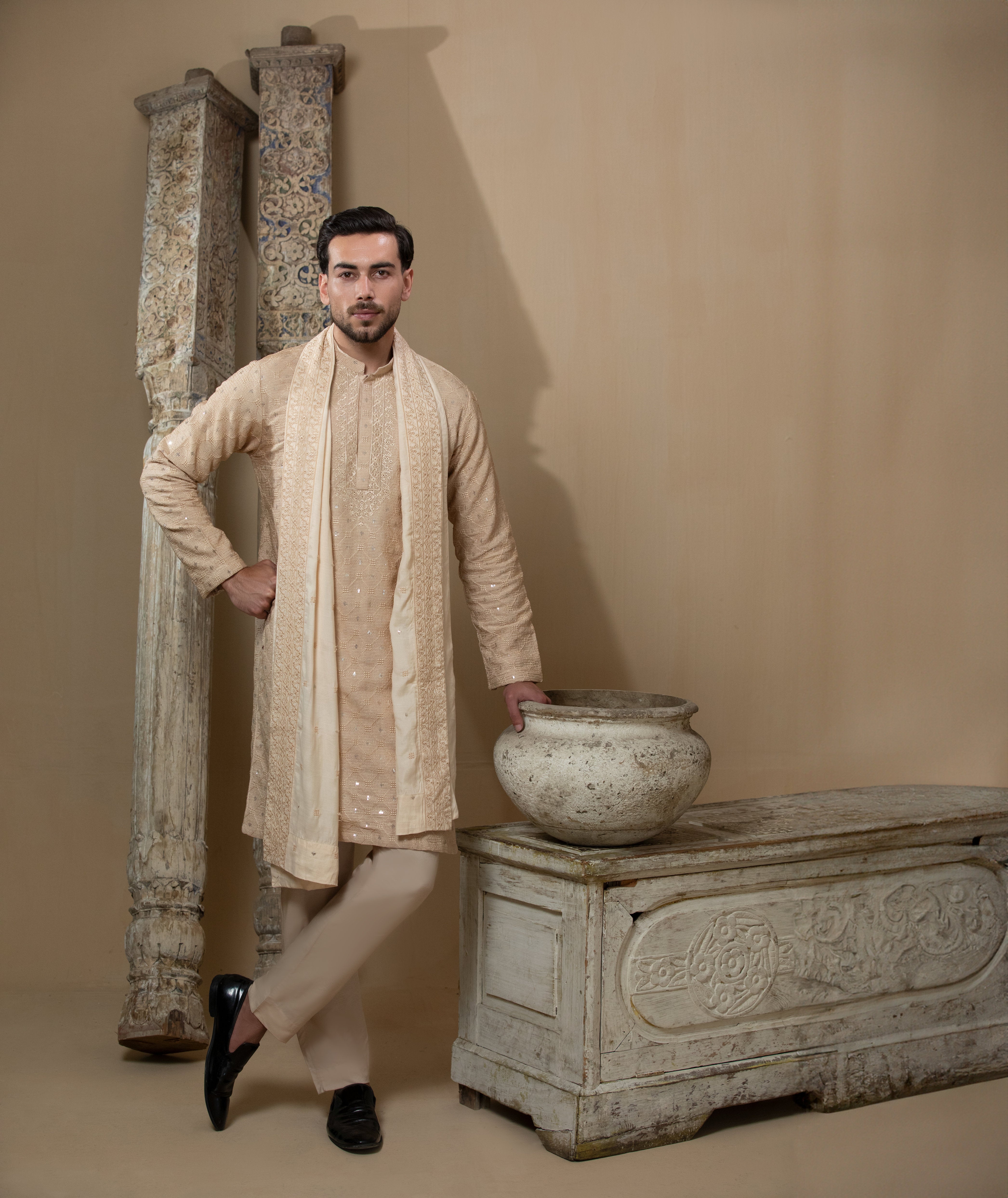 Man wearing Arsalan Iqbal's Peach & Gold Chikankari Kurta Pyjama with a cream stole draped over shoulders, styled with dark formal shoes against a warm beige backdrop