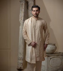 Man wearing Arsalan Iqbal's Peach & Gold Chikankari Kurta Pyjama with a cream stole draped over shoulders, styled with dark formal shoes against a warm beige backdrop