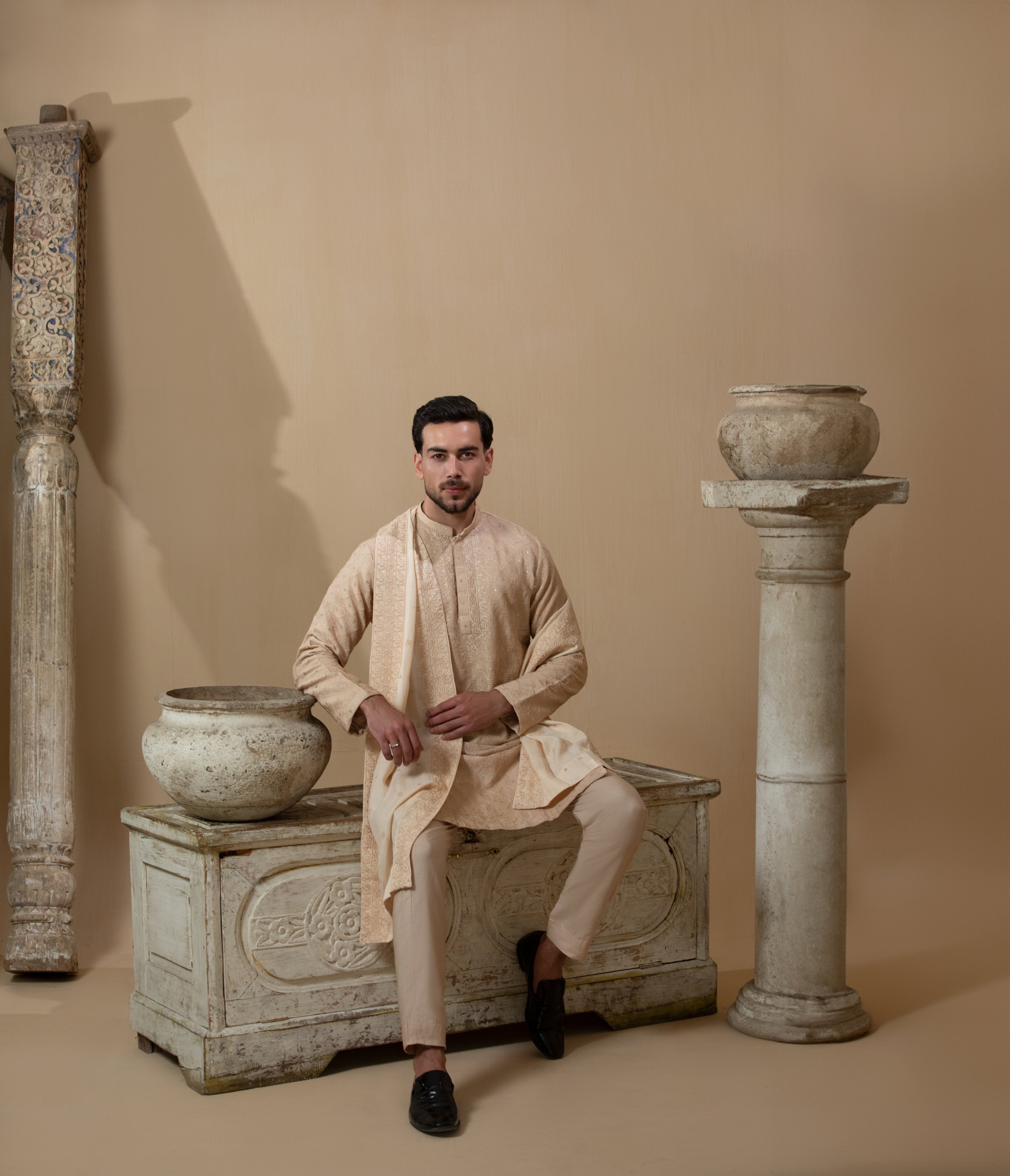 Man wearing Arsalan Iqbal's Peach & Gold Chikankari Kurta Pyjama with a cream stole draped over shoulders, styled with dark formal shoes against a warm beige backdrop