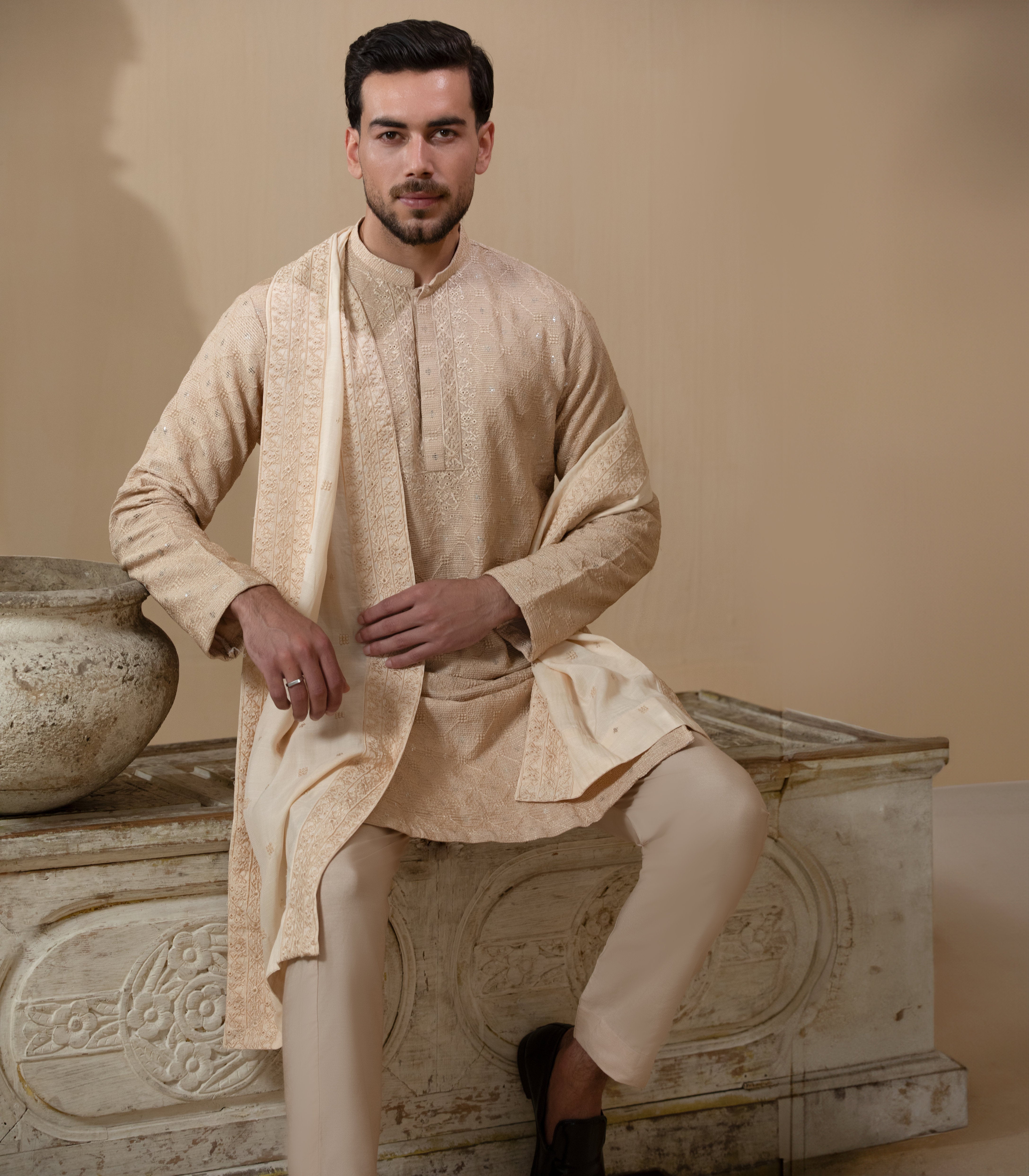 Man wearing Arsalan Iqbal's Peach & Gold Chikankari Kurta Pyjama with a cream stole draped over shoulders, styled with dark formal shoes against a warm beige backdrop