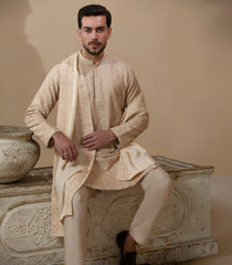 Man wearing Arsalan Iqbal's Peach & Gold Chikankari Kurta Pyjama with a cream stole draped over shoulders, styled with dark formal shoes against a warm beige backdrop