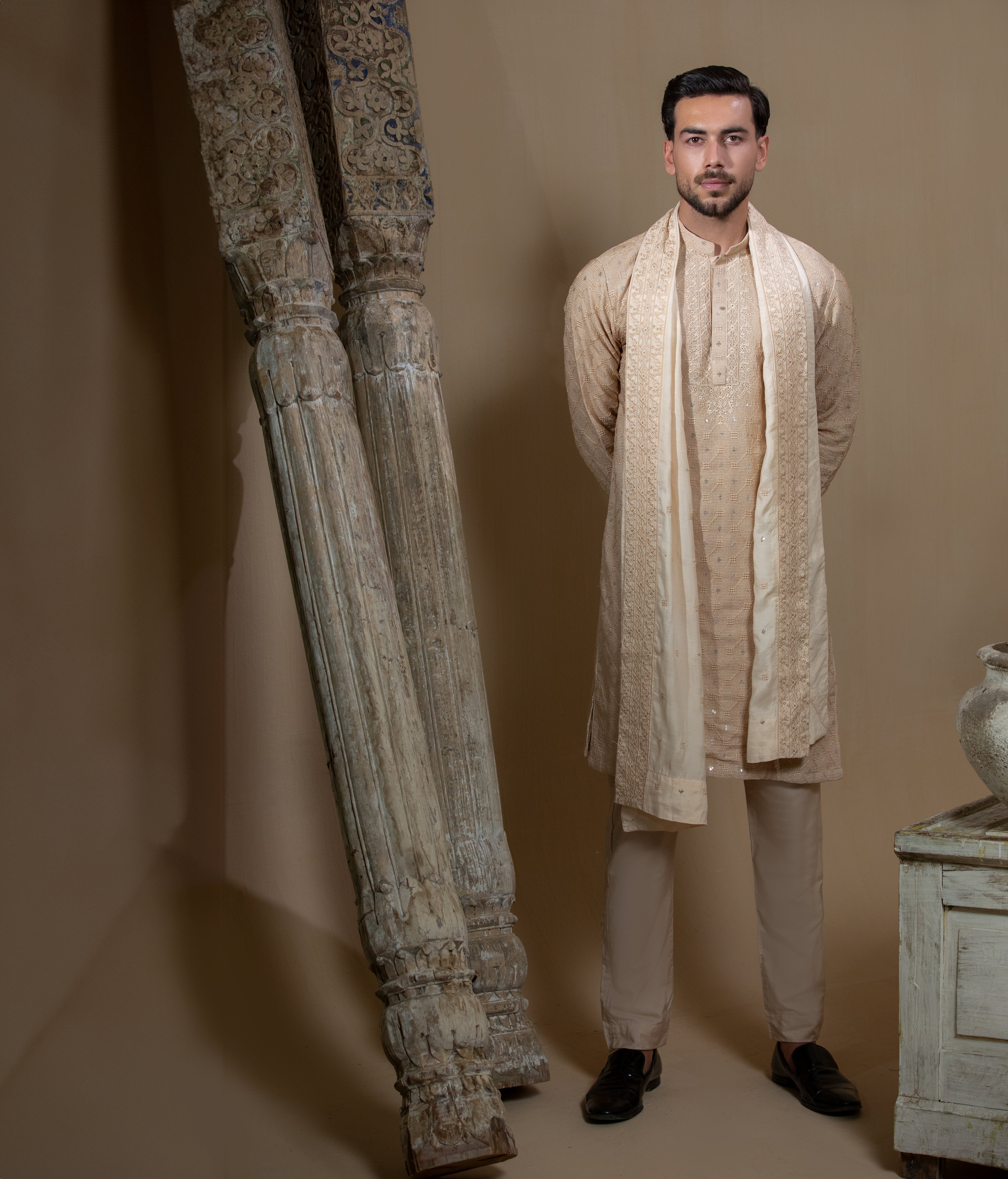 Man wearing Arsalan Iqbal's Peach & Gold Chikankari Kurta Pyjama with a cream stole draped over shoulders, styled with dark formal shoes against a warm beige backdrop