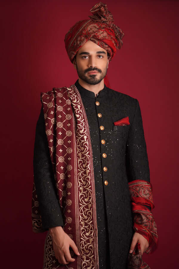 Waist-up portrait of an Arsalan Iqbal luxury black sherwani featuring gold buttons and a red silk pocket square, styled with a grand maroon silk turban and an intricately embroidered maroon shawl with golden floral motifs.