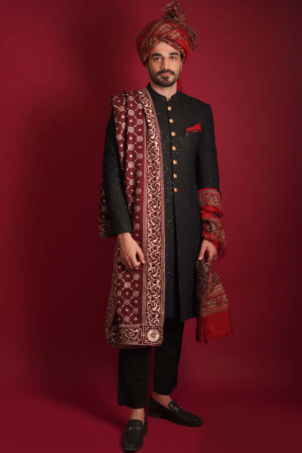 Full-length portrait of Arsalan Iqbal luxury black sherwani featuring gold buttons, paired with a matching maroon silk turban and an intricately embroidered maroon silk shawl with gold motifs, styled with black trousers and loafers.