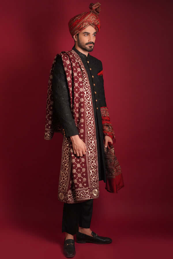 Side profile of Arsalan Iqbal luxury black sherwani with gold buttons, paired with a matching maroon silk turban and a heavily embroidered maroon silk shawl with golden motifs, styled with black trousers and loafers.