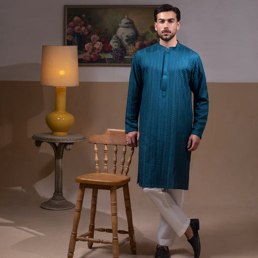 Arsalan Iqbal Teal Blue Chikankari Kurta Pyjama  man wearing a rich teal blue full-sleeve kurta with subtle all-over chikankari embroidery, mandarin collar with button placket, paired with white straight-cut pyjama, posed confidently with one hand on a wooden chair, styled in a warm classic indoor setting with a yellow vase and floral painting in the background