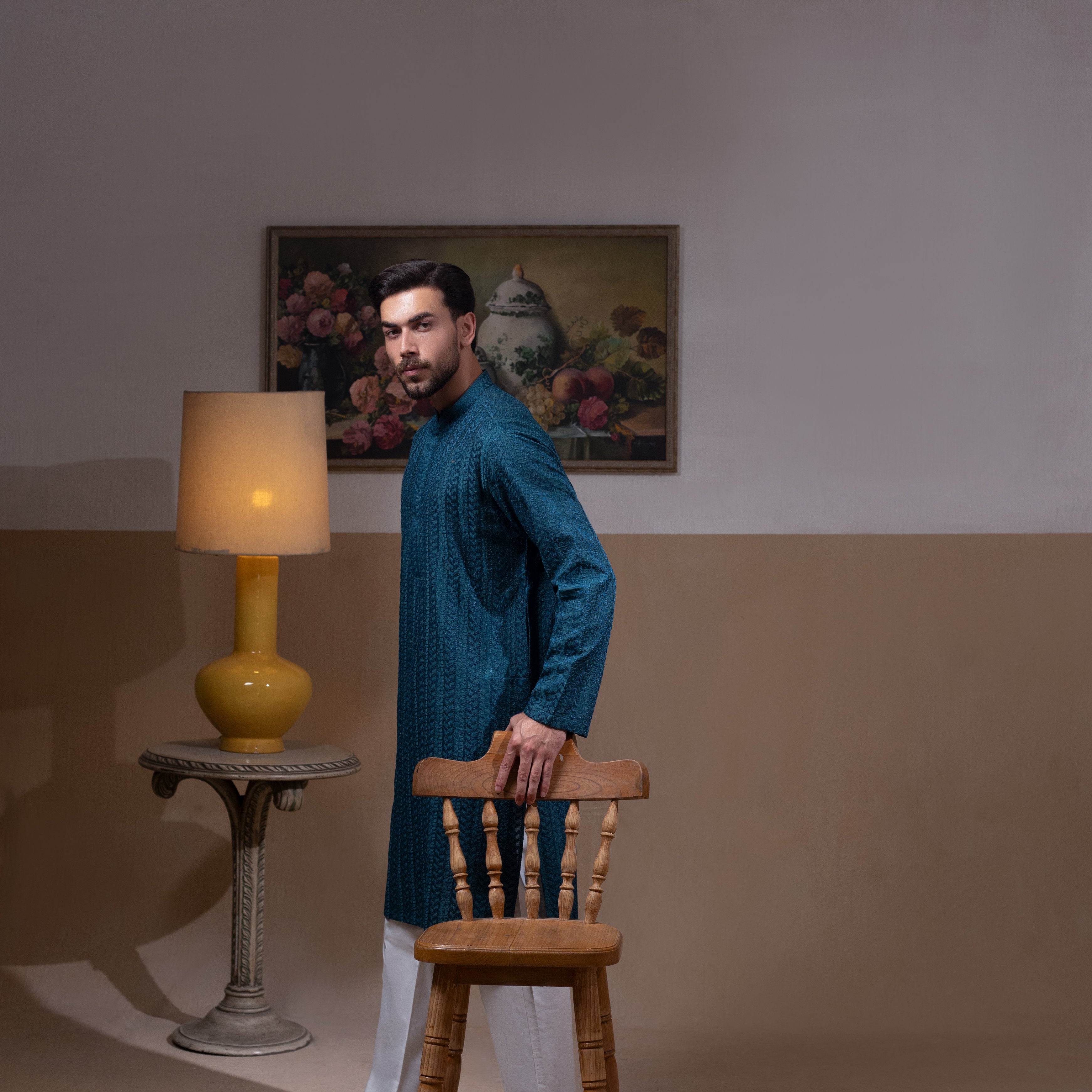 Arsalan Iqbal Teal Blue Chikankari Kurta Pyjama man wearing a rich teal blue full-sleeve kurta with subtle all-over chikankari embroidery, mandarin collar with button placket, paired with white straight-cut pyjama, posed confidently with one hand on a wooden chair, styled in a warm classic indoor setting with a yellow vase and floral painting in the background