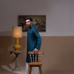 Arsalan Iqbal Teal Blue Chikankari Kurta Pyjama man wearing a rich teal blue full-sleeve kurta with subtle all-over chikankari embroidery, mandarin collar with button placket, paired with white straight-cut pyjama, posed confidently with one hand on a wooden chair, styled in a warm classic indoor setting with a yellow vase and floral painting in the background