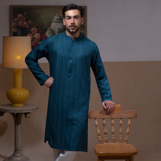 Arsalan Iqbal Teal Blue Chikankari Kurta Pyjama  man wearing a rich teal blue full-sleeve kurta with subtle all-over chikankari embroidery, mandarin collar with button placket, paired with white straight-cut pyjama, posed confidently with one hand on a wooden chair, styled in a warm classic indoor setting with a yellow vase and floral painting in the background