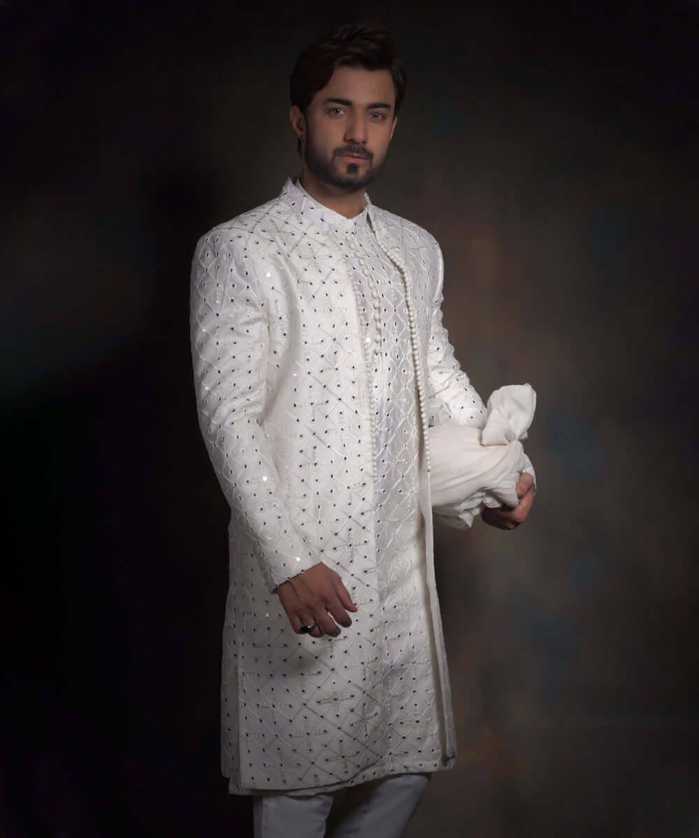 Portrait of Arsalan Iqbal snow white luxury sherwani for men, featuring geometric mirror-work, tonal embroidery, and a matching white turban with a row of decorative fabric buttons.