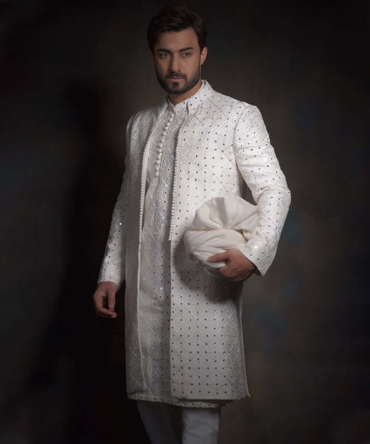 Side angle view of Arsalan Iqbal snow white luxury sherwani for men, featuring a decorative row of fabric buttons, intricate geometric mirror-work, and a matching white turban.