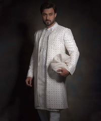 Side angle view of Arsalan Iqbal snow white luxury sherwani for men, featuring a decorative row of fabric buttons, intricate geometric mirror-work, and a matching white turban.