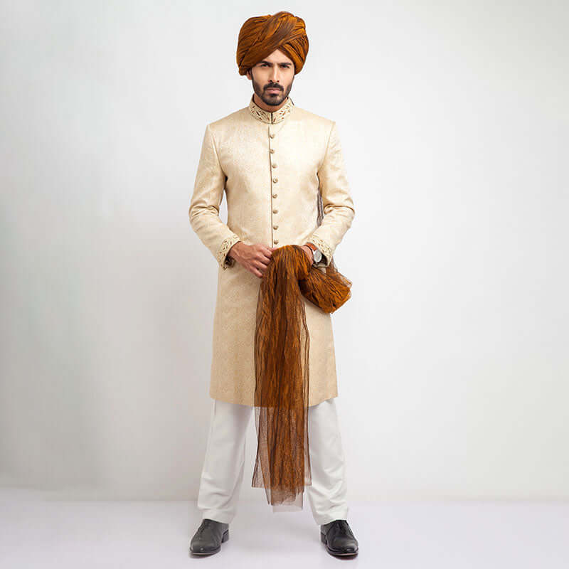 Full-length studio shot of a man in an Arsalan Iqbal black textured sherwani with a red pocket square, maroon turban, and white trousers.