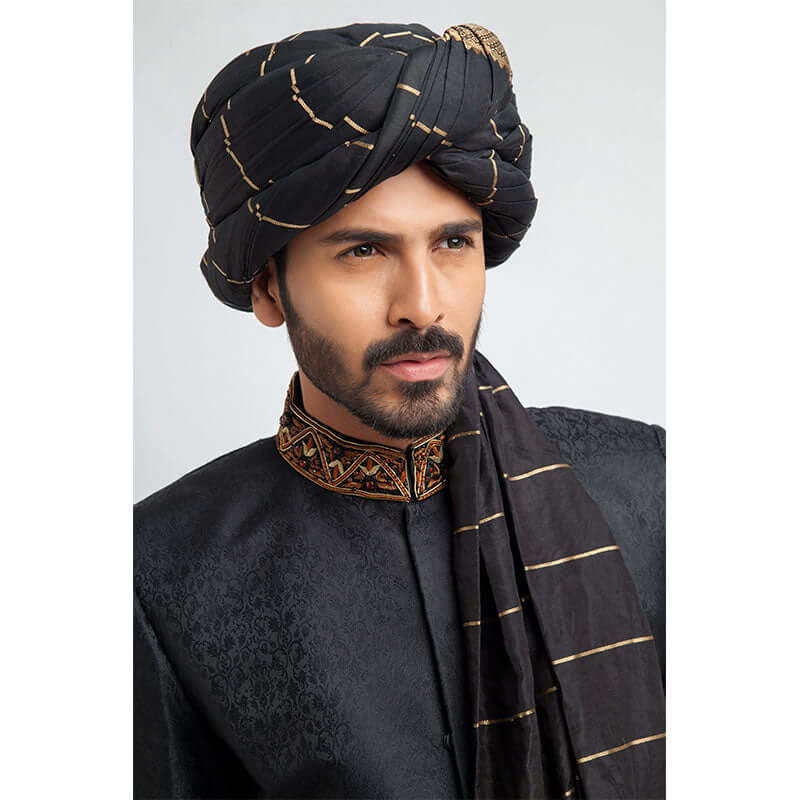 Close-up headshot of an Arsalan Iqbal black jacquard sherwani featuring an intricately embroidered copper and bronze mandarin collar and a black silk turban with gold horizontal stripes.