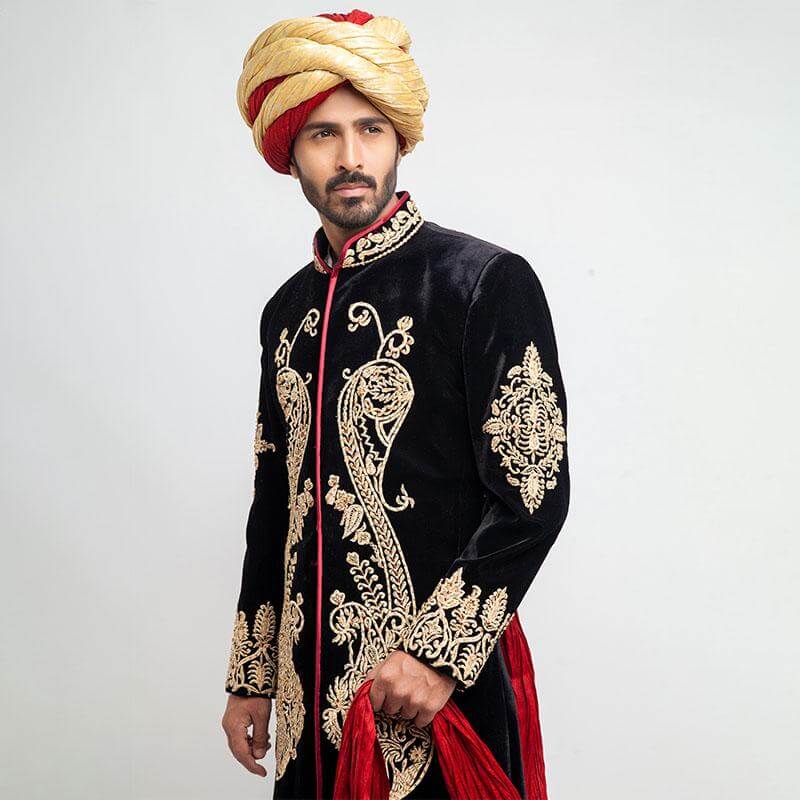 Three-quarter portrait of a man wearing an Arsalan Iqbal black velvet sherwani with intricate gold paisley chest embroidery and a gold and red dual-tone turban.