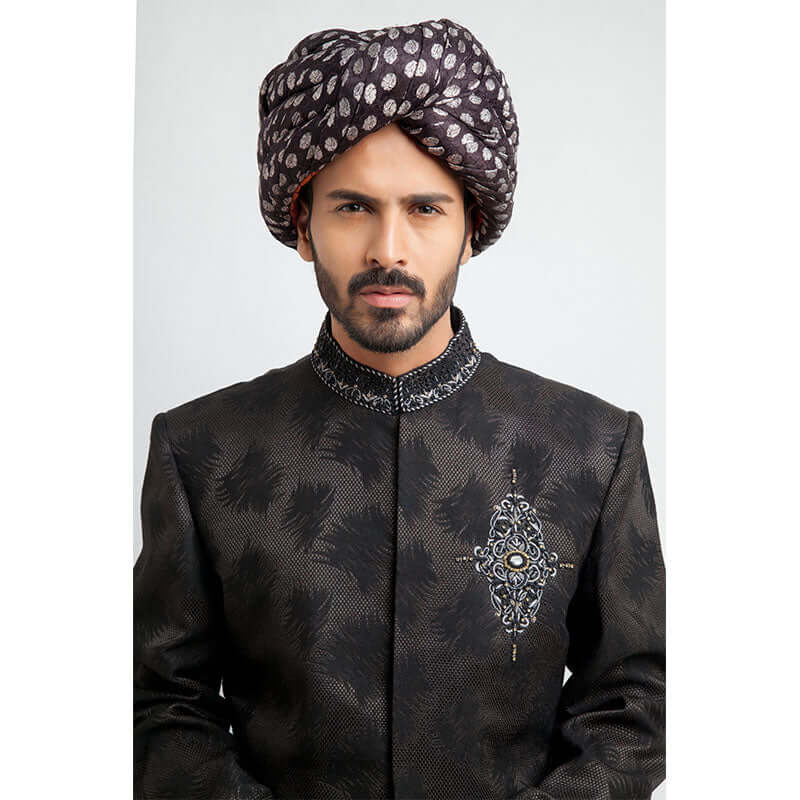 Close-up headshot of an Arsalan Iqbal charcoal grey jacquard sherwani highlighting the intricate silver and black embroidered chest emblem and a silk polka dot turban.