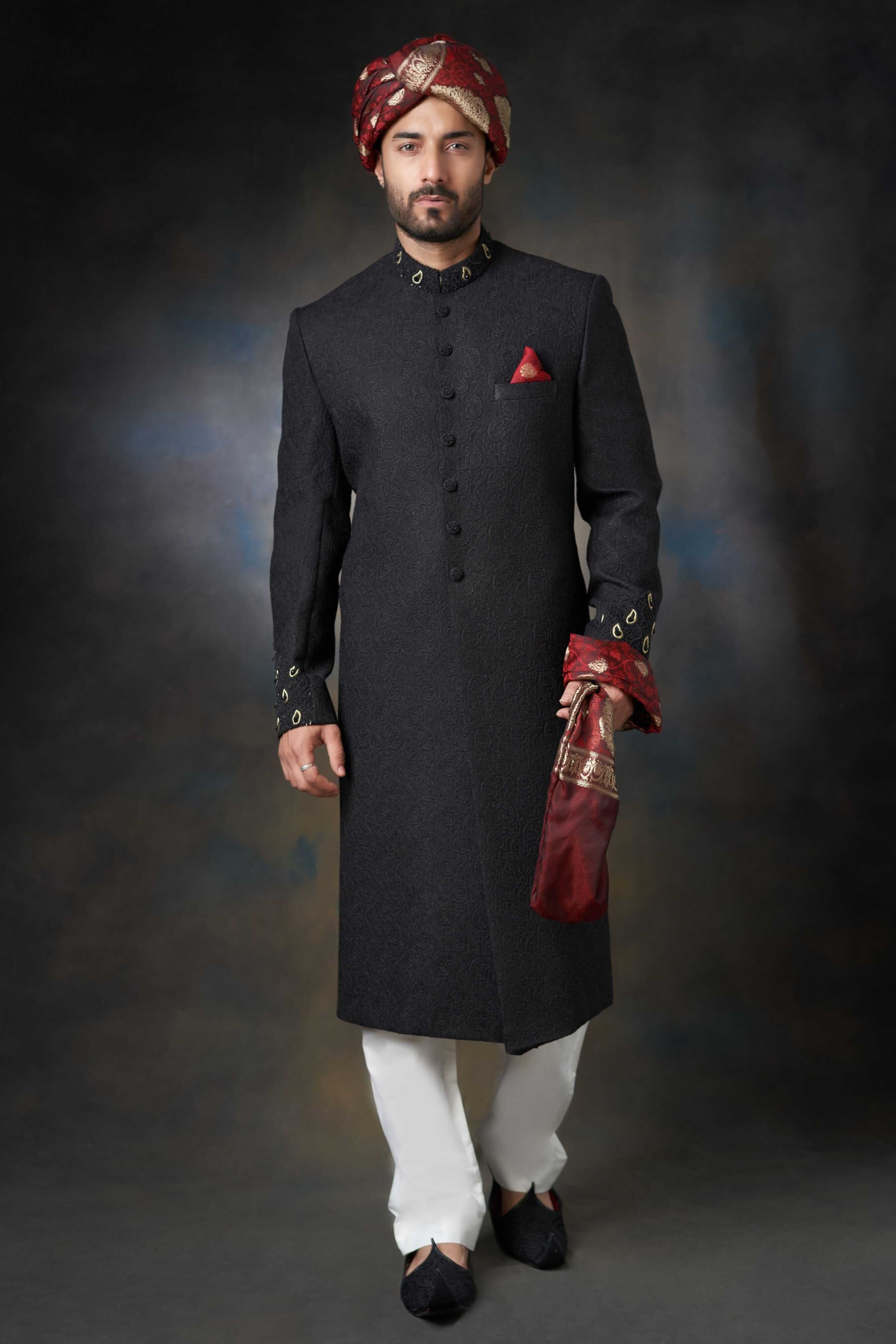 A front-view portrait of a man in a black Arsalan Iqbal embroidered sherwani, featuring a maroon pocket square, maroon and gold turban, and white trousers.