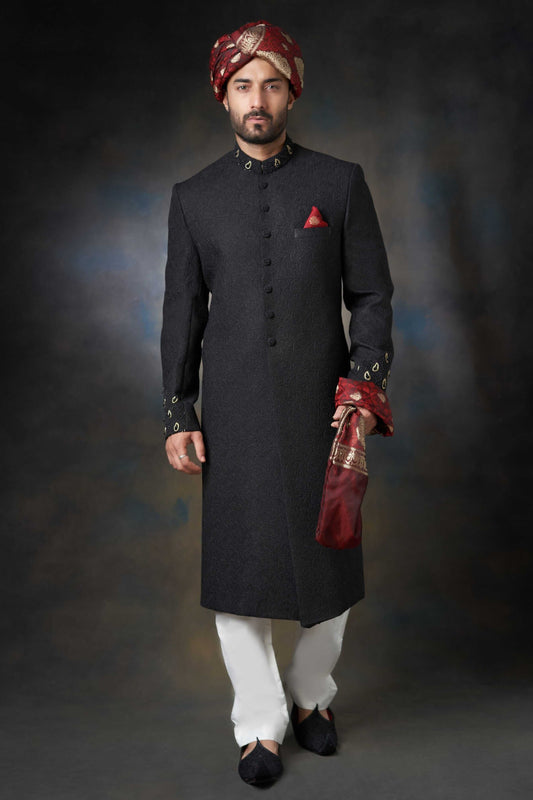 A front-view portrait of a man in a black Arsalan Iqbal embroidered sherwani, featuring a maroon pocket square, maroon and gold turban, and white trousers.