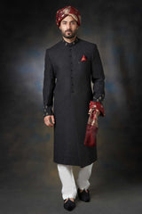 A front-view portrait of a man in a black Arsalan Iqbal embroidered sherwani, featuring a maroon pocket square, maroon and gold turban, and white trousers.