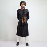 Full-length studio shot of a man in an Arsalan Iqbal black jacquard sherwani with an embroidered copper collar, paired with a black and gold striped turban and matching draped shawl.