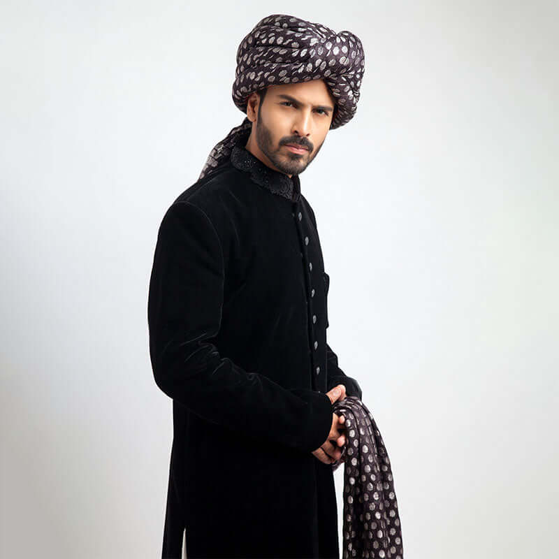Full-length studio view of a man in an Arsalan Iqbal black velvet sherwani, paired with a silver polka dot turban and matching shawl.