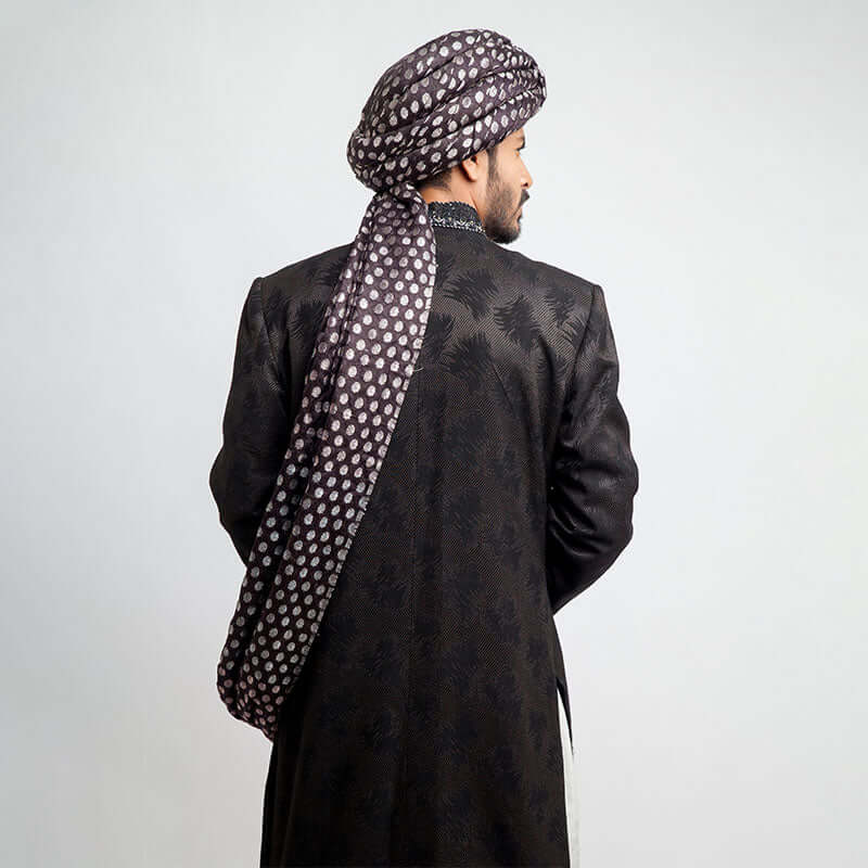 Rear view of an Arsalan Iqbal charcoal grey jacquard sherwani highlighting the tailored fit and the draped tail of a silver polka dot silk turban.