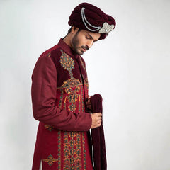 A three-quarter studio portrait of a man in an Arsalan Iqbal cream jamawar sherwani with a gold embroidered mandarin collar, paired with a vibrant red and gold polka dot silk turban and matching shawl.