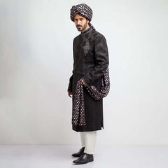 Full-length studio shot of a man in an Arsalan Iqbal charcoal grey jacquard sherwani featuring a metallic embroidered emblem on the chest, paired with a silver and charcoal polka dot turban and matching shawl.