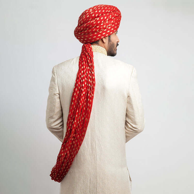 Rear view of an Arsalan Iqbal cream jamawar sherwani showing the tailored fit and the long, draped tail of a vibrant red and gold polka dot silk turban.