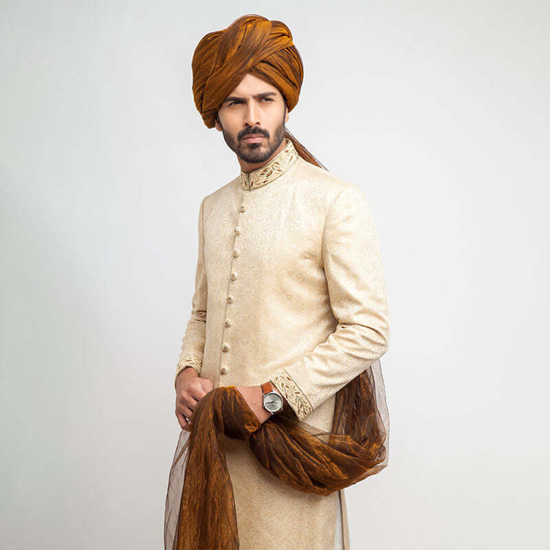 Full-length studio shot of a man in an Arsalan Iqbal gold jamawar sherwani with embroidered collar and cuffs, paired with a mustard yellow pleated turban, matching shawl, and white trousers.
