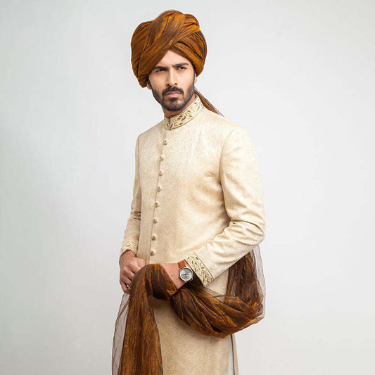 Full-length studio shot of a man in an Arsalan Iqbal gold jamawar sherwani with embroidered collar and cuffs, paired with a mustard yellow pleated turban, matching shawl, and white trousers.