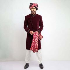 Full-length studio shot of a man in an Arsalan Iqbal maroon velvet sherwani with an asymmetrical button closure, paired with a pink and gold patterned brocade turban and matching shawl.