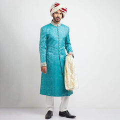 Full-length studio shot of a man in an Arsalan Iqbal teal jamawar sherwani with an all-over paisley pattern, featuring a silver embroidered mandarin collar and paired with a white and maroon striped brocade turban.