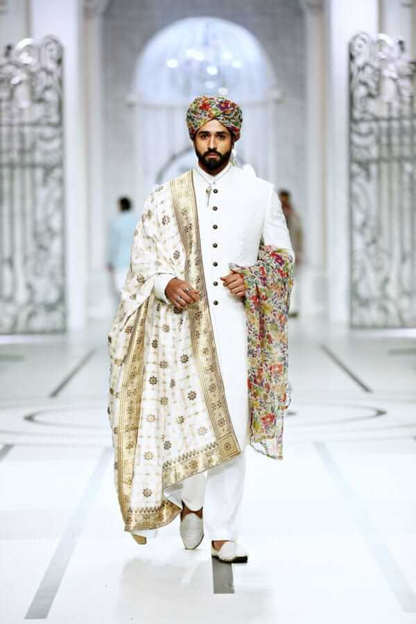 Full-length studio shot of a man in an Arsalan Iqbal black textured sherwani with a red pocket square, maroon turban, and white trousers.